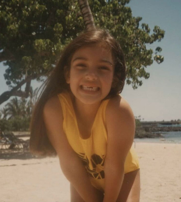 Young Kim Kardashian childhood photo smiling on a sunny beach, wearing a yellow swimsuit, with trees in the background.