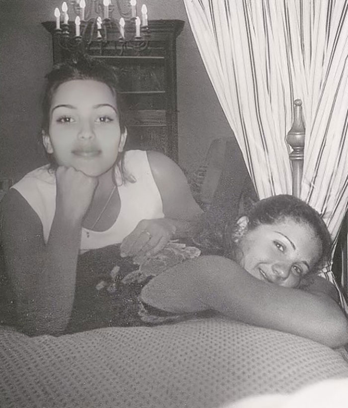 Young Kim Kardashian smiling with a friend, laying on a bed, under a chandelier.