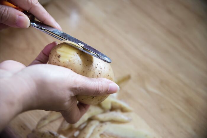 Left-handed person peeling a potato with a knife on a wooden surface.