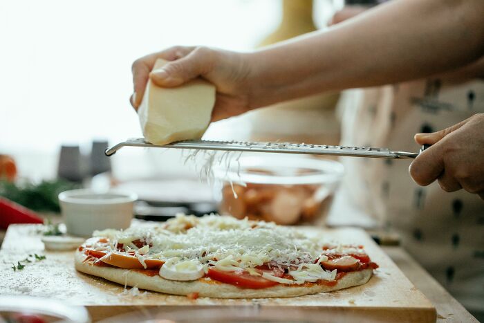 Left-handed person grating cheese onto a homemade pizza, highlighting everyday struggles in cooking tasks.