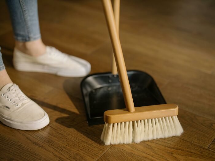 A left-handed person struggling with a broom and dustpan on a wooden floor, wearing white sneakers.