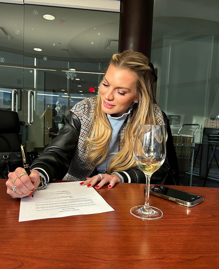 Reality TV star writing at a table, with a phone and wine glass nearby.