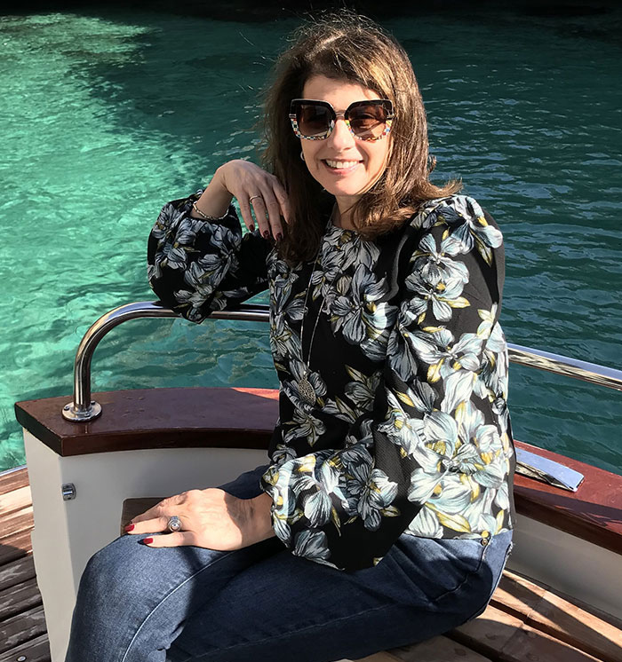 Luigi Mangione mother in floral blouse and sunglasses sitting by clear blue water, enjoying a sunny day outdoors.