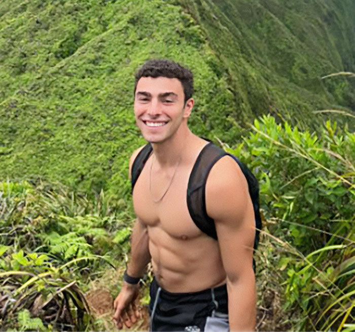 Luigi Mangione hiking shirtless with a backpack, smiling in lush green mountainous landscape.