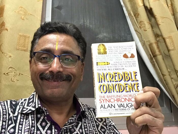 Man holding a book titled "Incredible Coincidence," smiling indoors, highlighting insane coincidences.