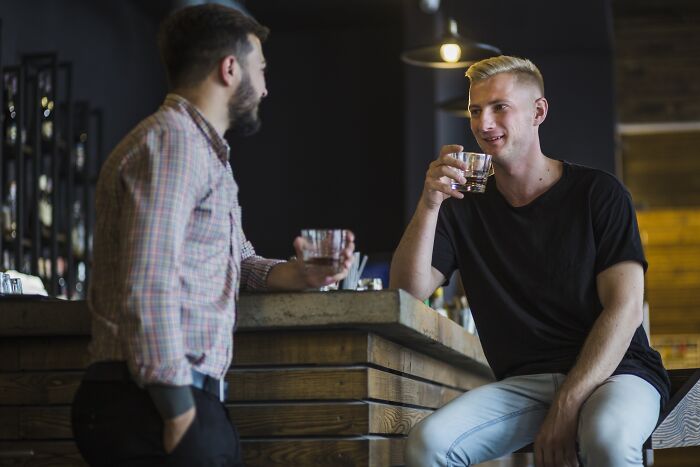 Two men chatting at a wooden bar, both holding glasses, experiencing an unexpected coincidence.