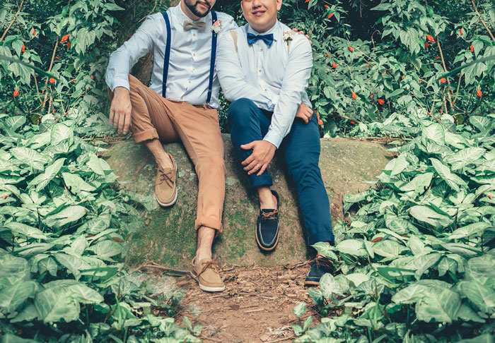 Two people sitting on a rock in a forest, dressed in matching shirts and bow ties, surrounded by greenery.