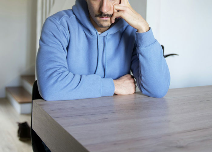 Man in a blue hoodie sitting at a table, appearing deep in thought about refusing to babysit.