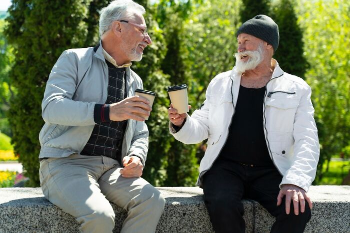 Two older men sitting on a bench, smiling and holding coffee cups, sharing secrets in a sunny park setting.