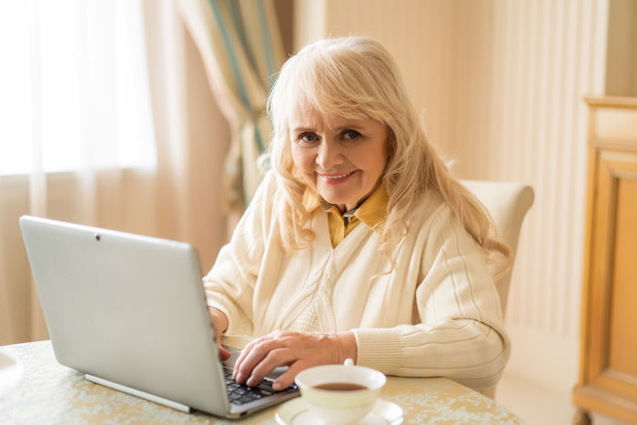 Elderly woman smiling at laptop in a cozy room, related to being mocked for her name "Dove" by family members.