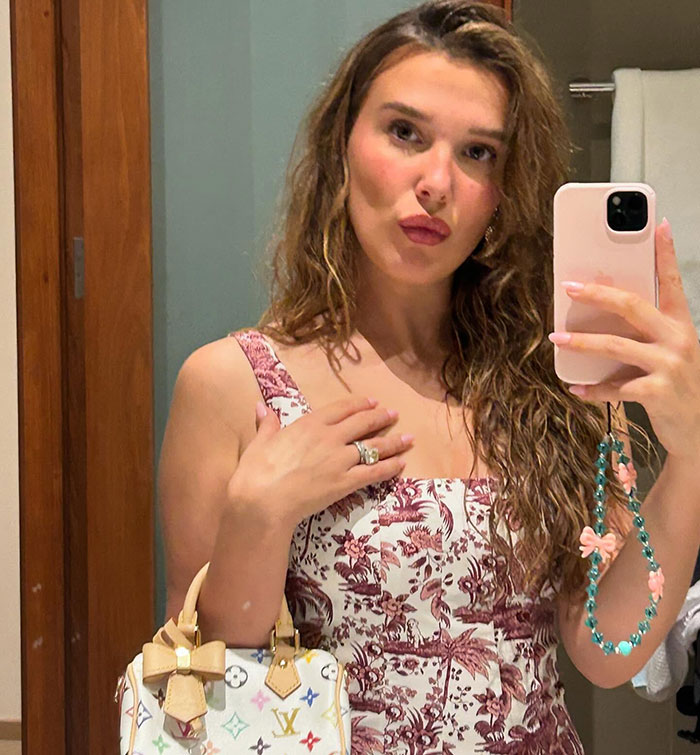 Young woman taking a mirror selfie, wearing a patterned dress and holding a designer bag, looks older.