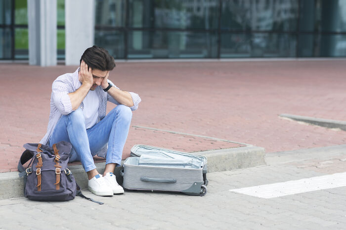 Traveler sitting on a curb, looking stressed with an open suitcase and backpack nearby, highlighting cringeworthy situations.