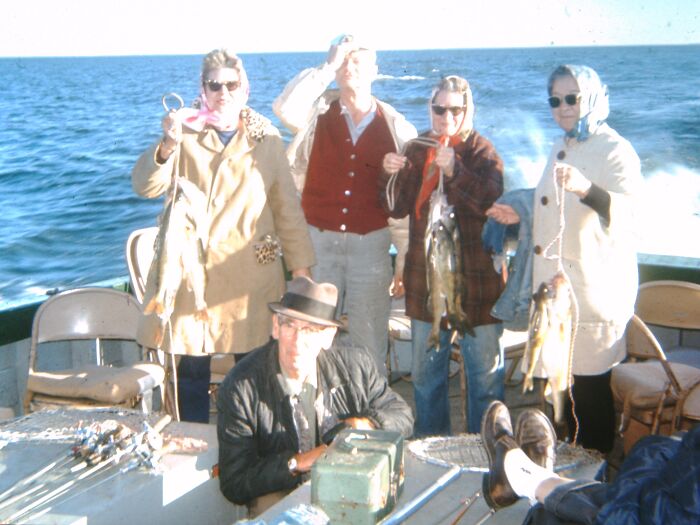 People in 60s fashion looks on a boat, holding fish, with ocean view in the background.