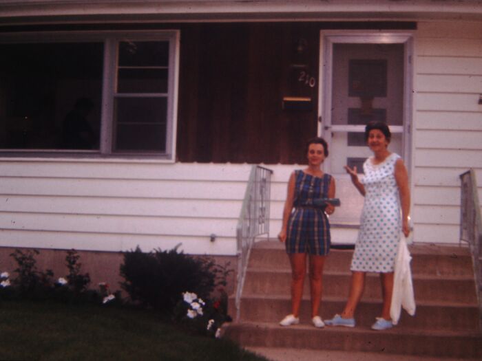 Two women in 60s fashion standing on house steps, one in a plaid dress and the other in a polka dot dress, smiling.