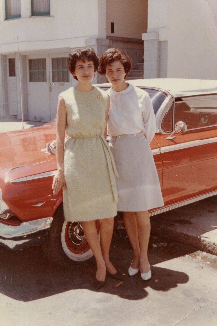 Two women in 60s fashion standing by a vintage red car, dressed in elegant dresses and classic hairstyles.