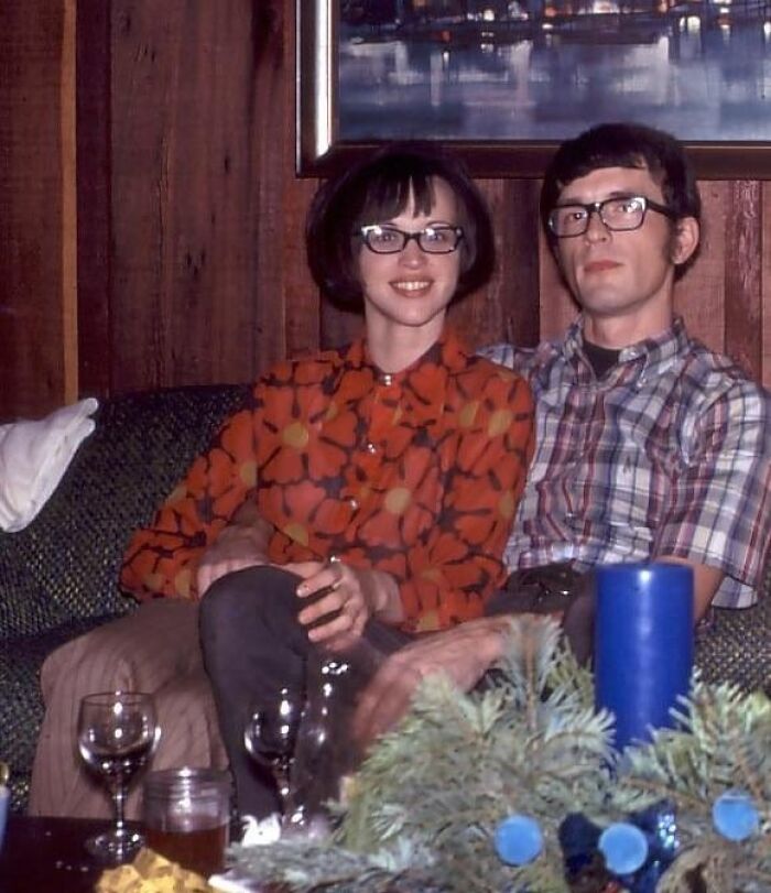 A couple sitting on a sofa in a room with 60s fashion looks, wearing glasses and vintage patterned clothing.