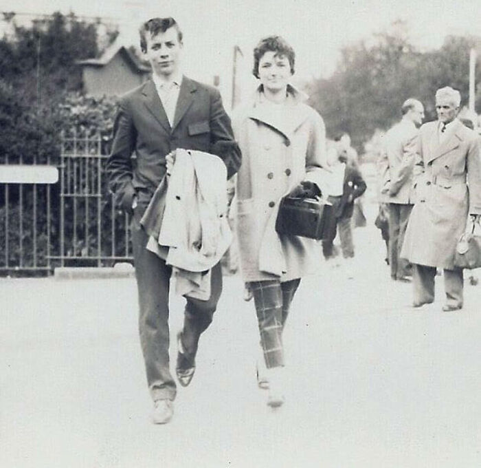 A couple showcasing 60s fashion looks, the man in a suit and the woman in a coat with plaid pants, walking outdoors.