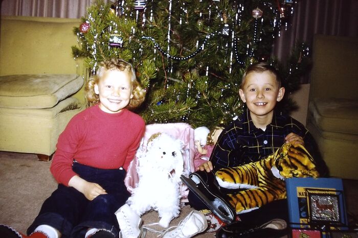 Two children in 60s fashion looks, sitting by a decorated Christmas tree with toys.