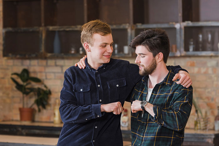 Two friends in casual shirts bumping elbows in a cozy kitchen setting, highlighting a moment of friendship.