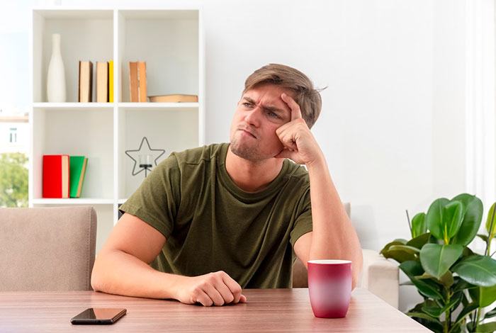 Young man in a green shirt sitting at a table, looking thoughtful and holding his forehead, phone nearby.