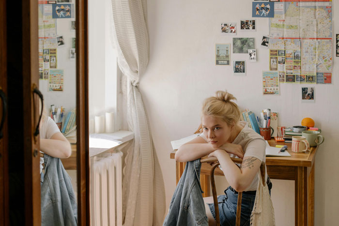 Young woman in a cozy room, contemplating, with photos and maps on the wall, capturing a mom-relationship-drama theme.