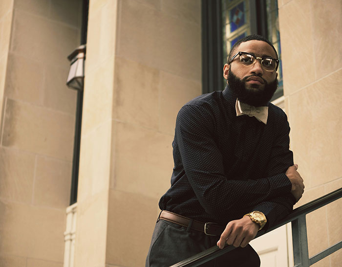 Man with a beard and glasses leaning on a staircase railing, wearing formal attire, challenging "born gifted" stereotypes.