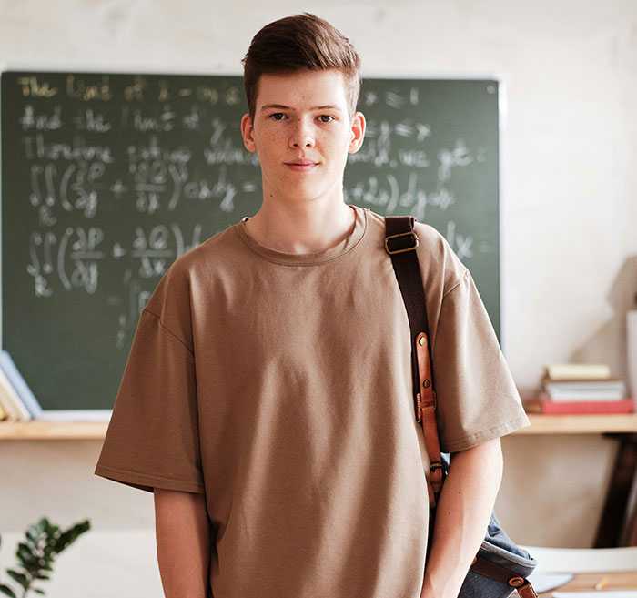 Young student with a backpack stands confidently in front of a chalkboard, challenging "Born Gifted" stereotypes.