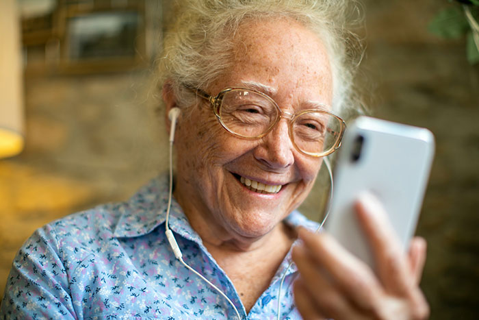 Elderly woman smiling, wearing glasses and earphones, using a smartphone, challenging stereotypes about technology.