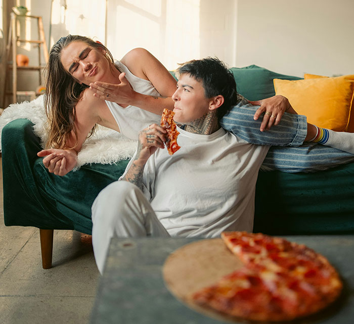 Two people on a couch playfully interacting, sharing pizza, humorously challenging stereotypes and misconceptions.