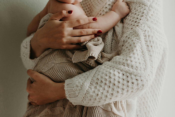 A woman in a white knit sweater holds a baby wrapped in a matching blanket.