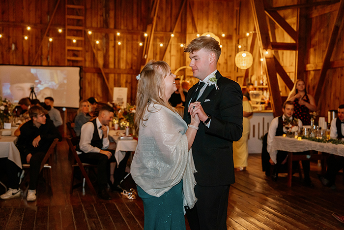 Groom dancing with mother at a summer wedding reception in a wooden barn venue, surrounded by guests.