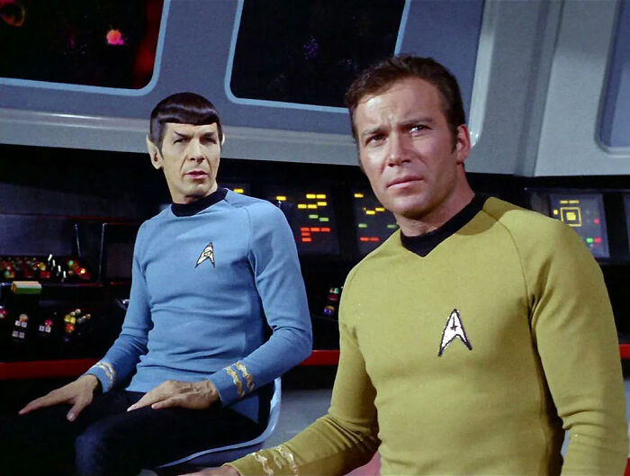 Two men in sci-fi uniforms on a spaceship bridge, highlighting movie portrayal issues.