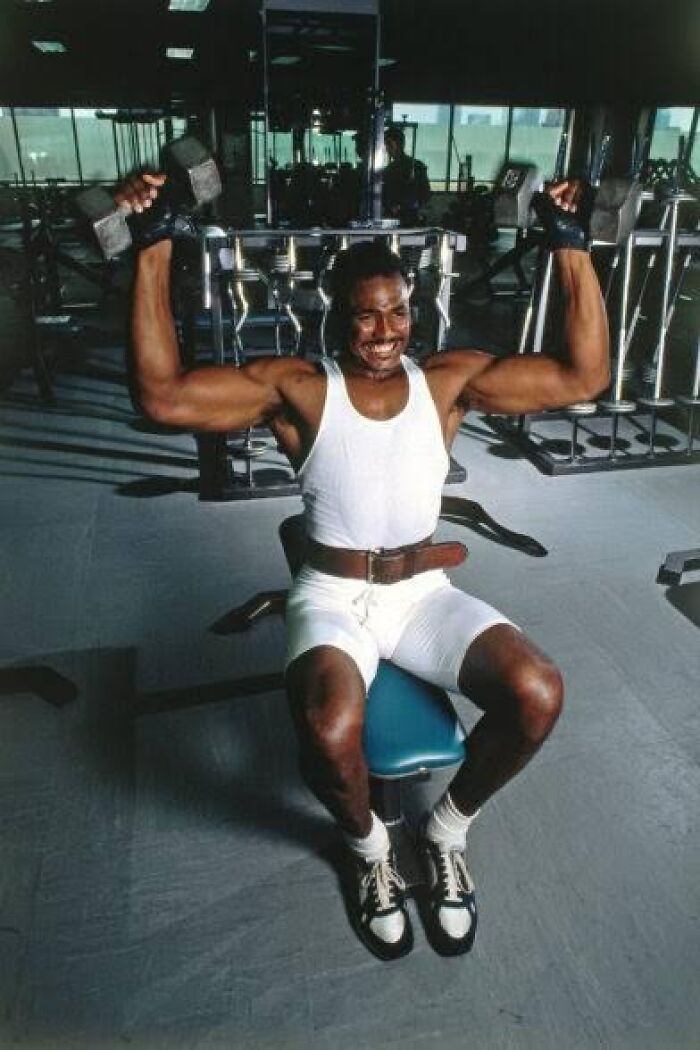 Man in white workout gear lifting dumbbells in a retro gym, showcasing 80s gym fitness style.