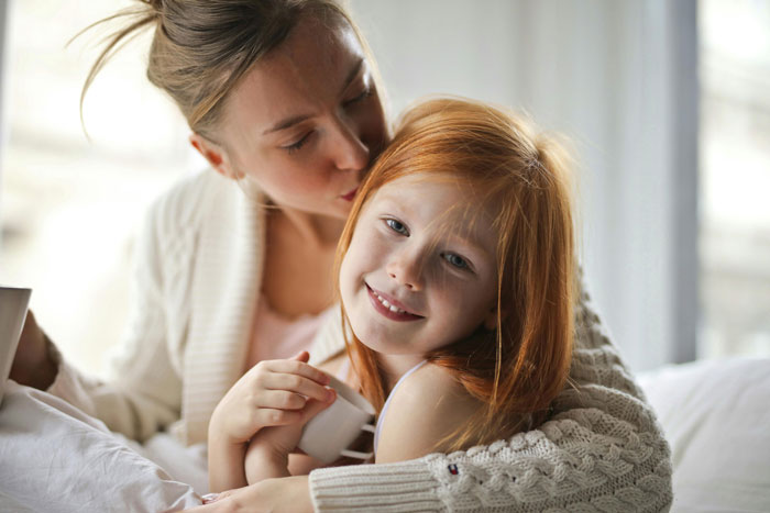 A mother and daughter share a tender moment on the bed, with the mother hugging and kissing her daughter's head.