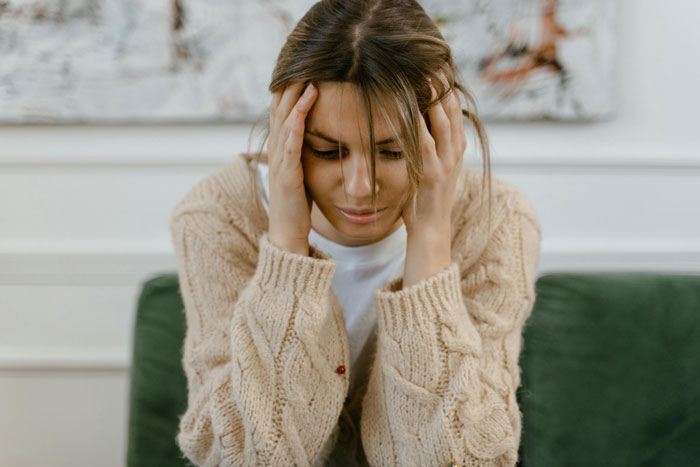 Woman in beige sweater sitting on a green couch, appearing distressed about a neighbour-daughter-husband-affair.