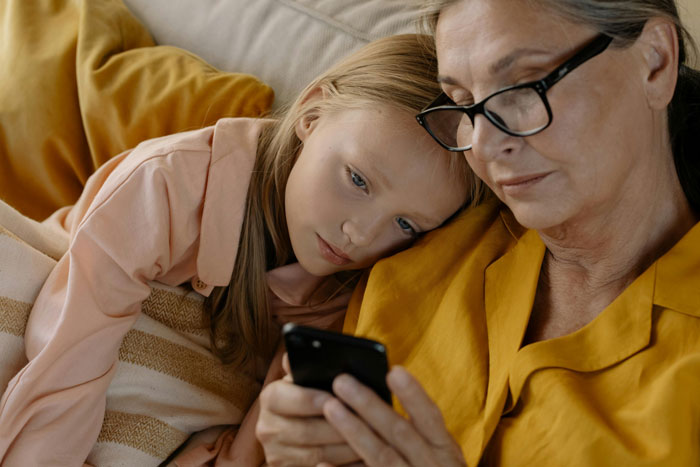 Elderly woman and young girl on a couch looking at a smartphone, the woman in glasses wearing a yellow shirt.