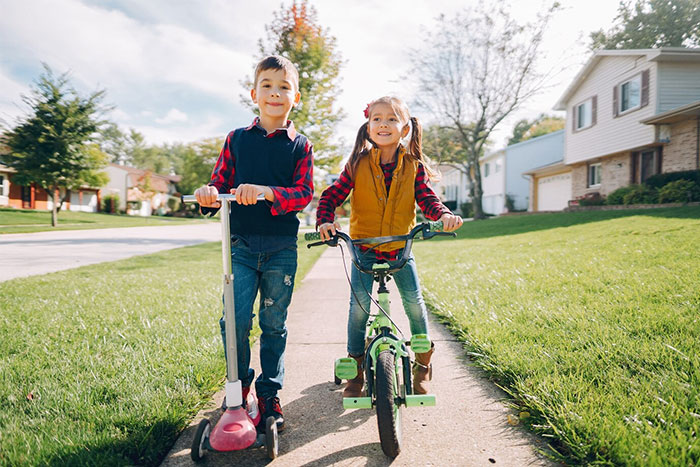 Kids playing outside on a scooter and bike, enjoying a sunny day, reminiscent of activities obvious 15 years ago.