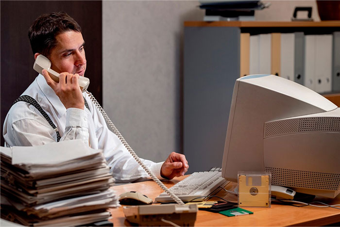 Man using a CRT monitor and landline phone, with floppy disks and paper stacks, reflecting old obvious technology.