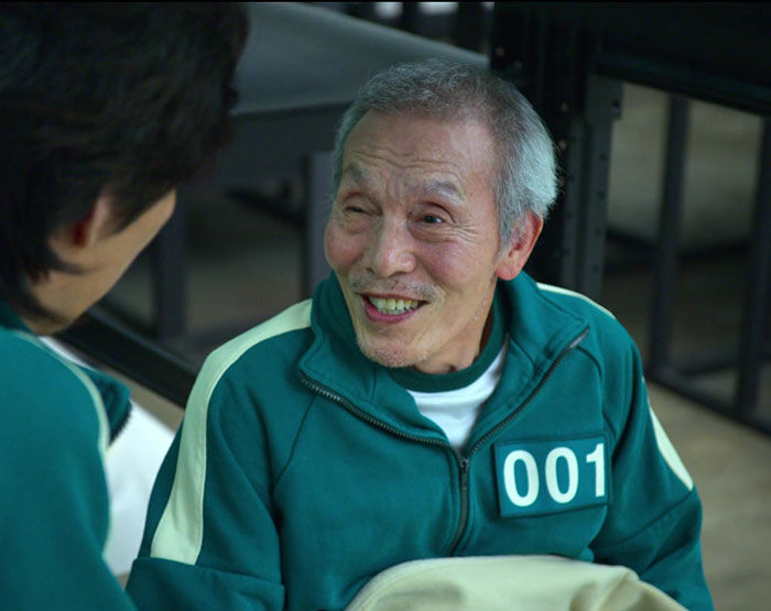 Senior man in uniform numbered 001 smiling, a scene from "Squid Game 2" amid cast controversy.