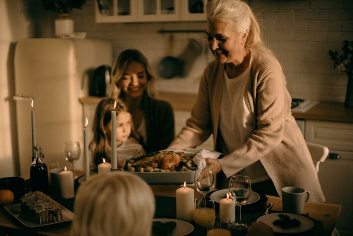 Elderly woman serving Christmas dinner to family, including children, in a warmly lit kitchen.