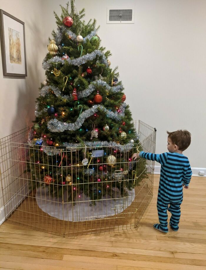 Child in striped pajamas looking at a Christmas tree behind a protective fence. Parenting-Genius-Tricks for holiday safety.