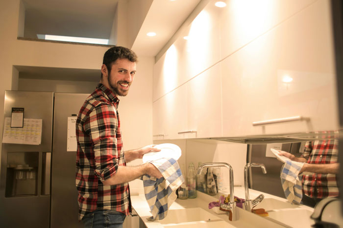 Man in a plaid shirt smiling while drying dishes in the kitchen, illustrating partner housework exchange.