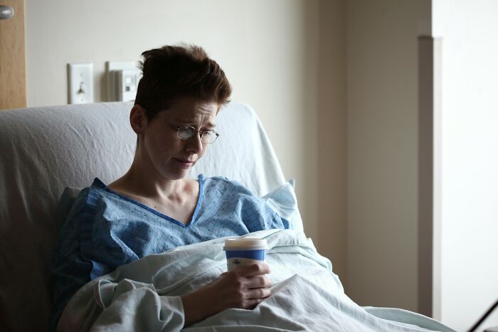 Person in a hospital bed, wearing glasses and holding a cup, embodying survival against all odds.