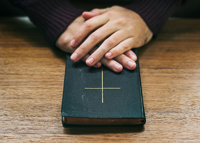 Hands resting on a closed book with a cross on the cover, symbolizing persistent Mormons' faith and learning.