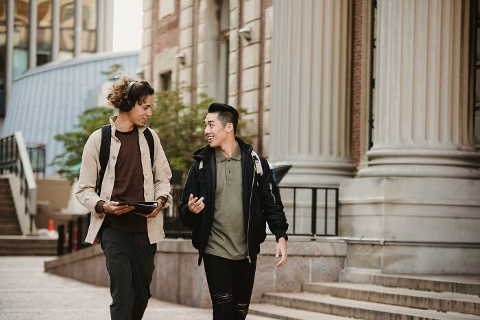 Two young men, dressed in casual clothing, walk and chat in front of a grand building, representing rich kids.