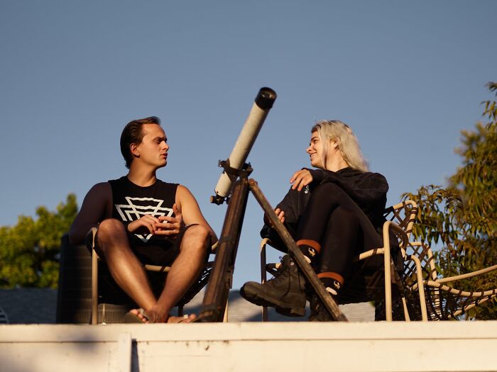 Two people casually talking on a rooftop, highlighting a dating mishap with a telescope between them, under clear skies.