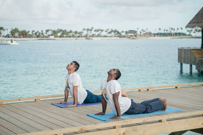 Two men doing yoga on a dock by the ocean, engaging in the WomenInMaleFields trend.