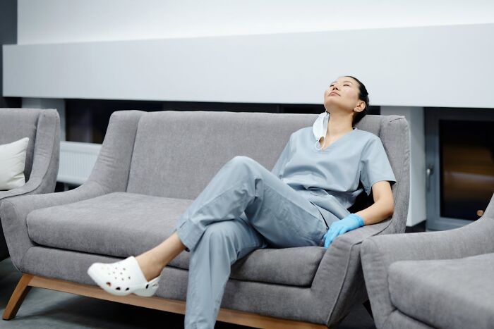 Person in scrubs looking exhausted, sitting on a gray couch with surgical gloves and crocs, representing unexpected job loss.