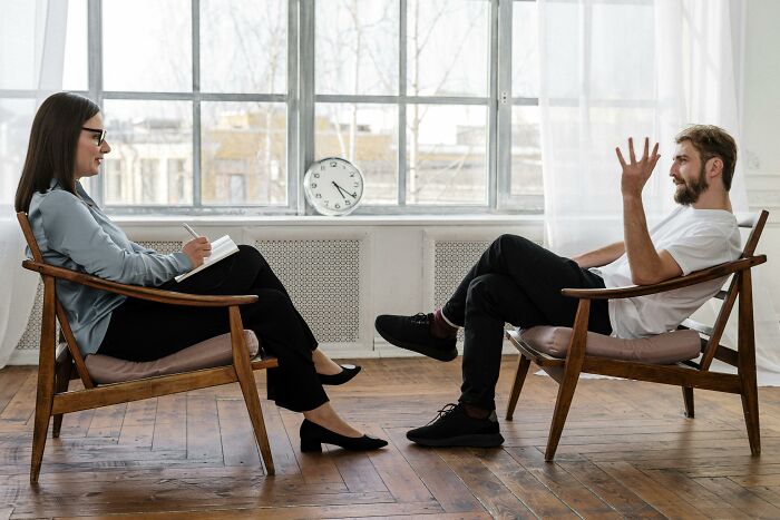 Two people in chairs by a window, engaged in a conversation, sharing secrets in a relaxed setting.