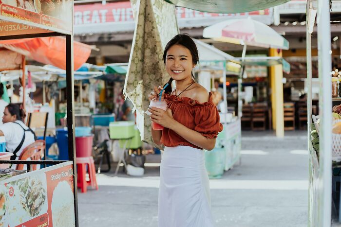 Woman smiling in a market, holding a drink, possibly experiencing an astonishing coincidence.